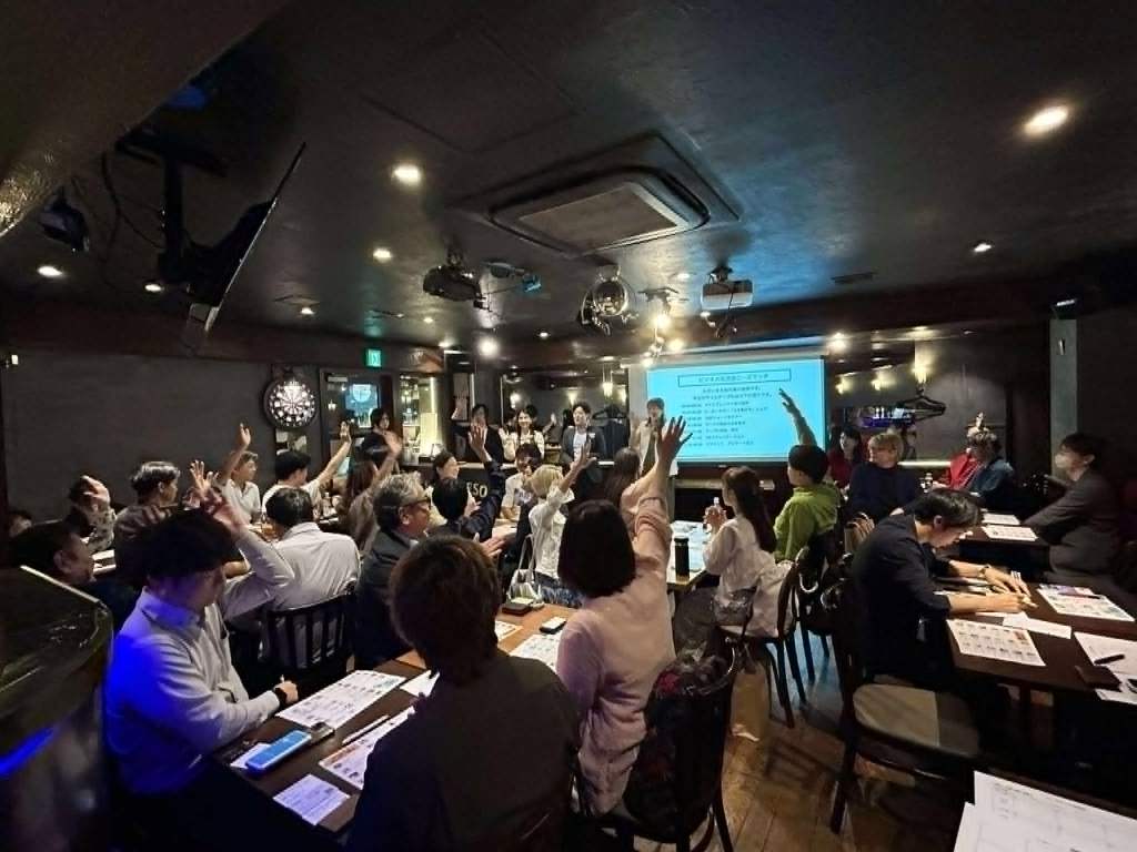 薄暗い飲食店の写真です。奥の壁にスクリーンがつるされて、何らかの画面が映っています。ほとんど満席に近いほどの人が着席しており、そのうちの多くが挙手をしています。スクリーンの横に3人の人が立って司会進行をしている様子が見えます。