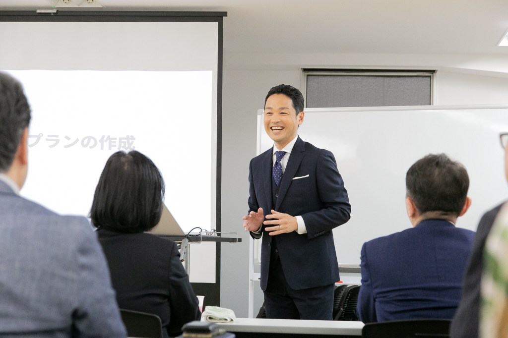 セミナーの様子を写した写真です。中央にスーツ姿の箱山さんが立ち、何か話しています。隣のスクリーンには白い背景に「プランの作成」と書かれた画面が映し出されています。手前に何人かの聴衆の後ろ姿が見え、いずれの人もジャケットを着用しています。