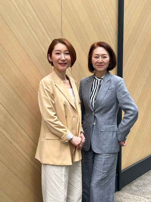ジャケットスタイルの2人の人が並んで立っている写真です。右にこのインタビューの話し手である鈴木さんが、グレーのスーツスタイルで写っています。左の人は薄橙色のジャケットに白っぽい色のズボンを身に着けています。